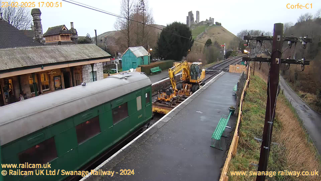 A view of Corfe Castle railway station in a rural setting. The foreground features a green train carriage on the left and a yellow construction vehicle positioned beside the track. There are benches along the platform, and a blue building in the background. The castle can be seen atop a hill in the background, surrounded by trees. The sky is overcast, indicating a cloudy day.