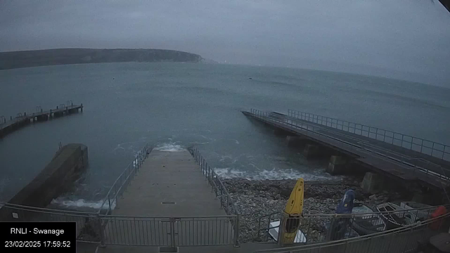 A cloudy coastal scene featuring a rocky shore and a calm sea. In the foreground, a boat ramp leads down to the water, bordered by a metal fence. To the right, several boats, including a yellow kayak and a blue one, are resting on the pebbled beach. In the distance, a sloping hill can be seen against the gray sky. The lighting is dim, suggesting late afternoon.