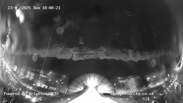 A nighttime webcam image showing a coastline. The view features ocean waves crashing onto the shore, illuminated by city lights in the background. The scene is slightly blurry due to water droplets on the lens. The date and time displayed at the top indicate it is Sunday, February 23, 2025, at 18:00:21. The bottom of the image contains the watermark "Powered By Brighton CCTV" and a website URL.