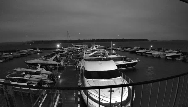 A nighttime image of a marina filled with several boats docked at slips. The scene is mostly dark with artificial lights illuminating parts of the marina and reflecting off the water. In the foreground, there is a large white boat that is more visible due to the lighting, while smaller boats are scattered throughout the marina in various orientations. In the background, there are distant landforms, likely an island or shoreline, silhouetted against the night sky. The overall atmosphere appears calm and serene.