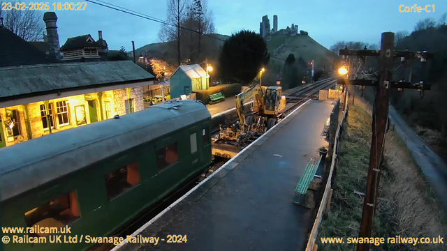 A view of a railway station during dusk, with a green train car partially visible on the left. In the foreground, there is construction equipment on the tracks and a platform with a shiny surface, likely from recent rain. To the right, there is a wooden bench and several street lamps casting a warm light. The background features a hill with castle ruins, partially obscured by trees, and several buildings with illuminated windows. A power pole stands on the far right, with visible wires overhead.