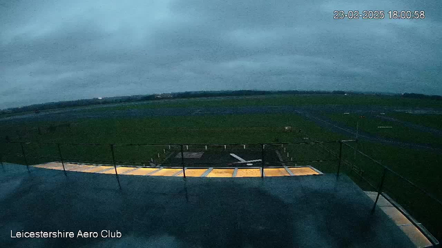 A view from a webcam situated on a tower at Leicestershire Aero Club. The scene shows a large, open green space, which is likely an airfield, under a gray, overcast sky. The ground is mostly flat with a bitumen runway visible extending into the distance. There are various markings on the runway, and a small white aircraft silhouette can be seen parked on the ground towards the center. The image is dimly lit, suggesting it is either dawn or dusk.