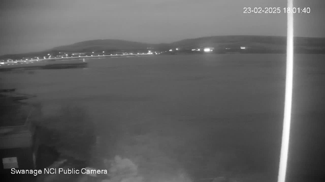 A black and white webcam image showing a calm body of water extending to the horizon. In the foreground, there is a dark area suggesting shoreline or land, and in the background, soft hills gently rise against the sky. Small lights are visible along the waterfront, twinkling faintly in the evening light, reflecting off the water's surface. The scene is tranquil with a sense of dusk falling. The image is time-stamped at 18:01 on February 23, 2025.