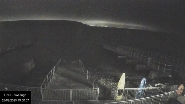 A dark scene captured by a webcam showing a coastal area at night. In the foreground, a fenced pathway leads towards the water, with a rocky shoreline visible along the bottom. Two kayaks, one yellow and one blue, are positioned next to the railings on the left. The background is dimly lit, with faint light on the horizon suggesting distant land or lights. The overall atmosphere is quiet and still.