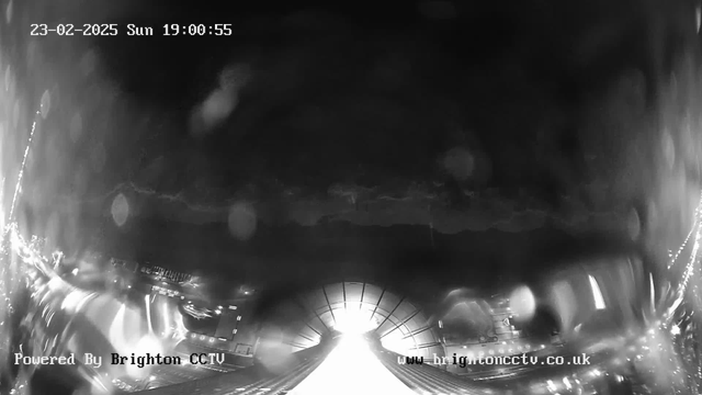 A black and white webcam image showing a view of a street or bridge illuminated by bright lights ahead. The foreground is dominated by a wide, brightly lit area that narrows towards the horizon. Blurry outlines of structures can be seen on both sides, possibly indicating buildings or vehicles. The sky is dark with hints of clouds. The date and time are displayed at the top of the image. A watermark indicating "Powered By Brighton CCTV" appears at the bottom left, with a web address on the bottom right.