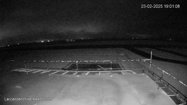 A nighttime view from a webcam at Leicester Aero Club. The scene is dark and shows an empty tarmac area with a railing in the foreground. There are faint lights visible in the distance, likely from a nearby settlement or airport. The ground surface appears flat and unoccupied. The date and time are displayed in the corner.