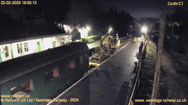 A nighttime view of a railway station platform. On the left, a green train car is partially visible, while on the right, construction equipment, including a yellow excavator, is in operation on the tracks. The platform is illuminated by several lights, and there are benches along the side. In the background, there are buildings with lit windows and some trees. The scene appears quiet with no people present.