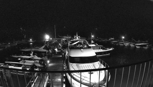 A dimly lit marina scene at night, featuring several boats lined up along a wooden dock. In the foreground, a large white boat with a smooth hull is prominently displayed. The background is filled with various smaller boats, and several bright lights are illuminating the area, reflecting off the water. The overall atmosphere is calm and serene, with the darkness surrounding the scene, highlighting the illuminated boats.