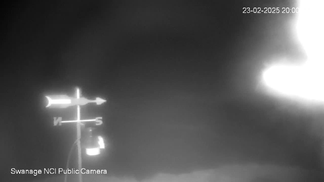 A black and white image from a webcam showing a directional weather vane with arrows pointing North and South. The background is dark and cloudy. In the lower part of the image, there is a light source visible, possibly a lamp. The top right corner displays a timestamp indicating the date and time as 23-02-2025 at 20:00. The lower left corner features a label identifying the camera as "Swanage NCI Public Camera."