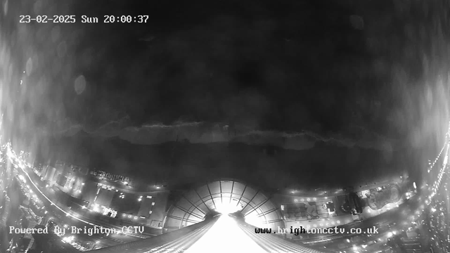 A black and white image from a webcam showing a cityscape at night. In the foreground, there is a bright light source at the center, possibly a large structure or building. The surrounding area features streets lined with lights indicating vehicles and buildings. The upper part of the image displays a dark sky with faint outlines, suggesting low clouds or distant hills. The timestamp in the upper left corner shows the date as February 23, 2025, and the time as 20:00:37. The bottom of the image includes a watermark with web address details.