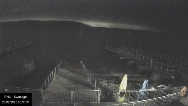 A dimly lit scene shows a coastal area with a rocky shoreline. In the foreground, there is a walkway surrounded by a fence, leading to a pier that extends into the water. On the ground, there are two kayaks, one yellow and one blue, positioned beside the walkway. In the background, a dark hillside is visible, with faint lighting on the horizon indicating possible distant buildings or streetlights. The overall atmosphere is calm and quiet, with only natural features and equipment in view.