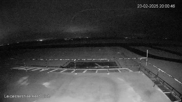 A dimly lit view from a webcam showing a runway or tarmac area, with a few scattered lights in the distance suggesting nearby buildings or structures. The foreground is mostly unmarked, with a railing visible along the edge, and a rectangular area is highlighted, possibly indicating a parking space or a boundary. The timestamp indicates it is night.