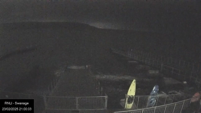 A webcam view at night showing a dark landscape with a faint outline of hills in the background. In the foreground, there are two kayaks resting against a railing: one is yellow and the other is blue. The surrounding area is mostly obscured by darkness, with some indistinct shapes of structures and a fence visible. The date and time display in the corner indicates it is February 23, 2025, at 21:00:03.