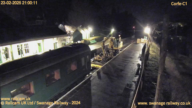 A nighttime scene at a railway platform, with low lighting. A green train is partially visible on the left side of the image. On the right, a construction vehicle is parked next to the track, with work equipment scattered nearby. There are lights illuminating the area and a building with windows can be seen in the background, creating a cozy but dim atmosphere. The ground appears wet, possibly due to recent rain.