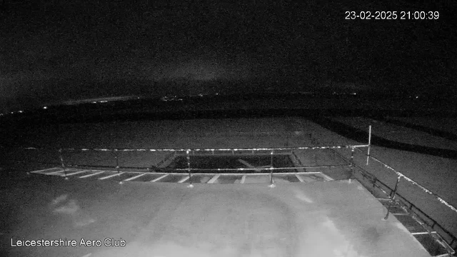 A dark, nighttime view from a webcam at Leicestershire Aero Club. In the foreground, there is a barrier or railing, and the ground appears flat and empty. The sky is mostly dark, with faint lights visible in the distance, suggesting the presence of towns or vehicles. The date and time are displayed in the top right corner.