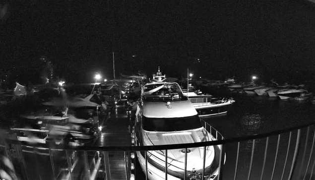 A nighttime image of a marina, featuring several boats docked along a wooden pier. The scene is illuminated by soft lights, reflecting off the water. The boats vary in size and shape, some with masts and others with covers. The overall atmosphere is calm and serene, showcasing the quiet of the night.