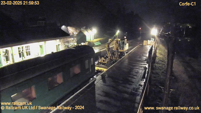 A dark image showing a railway station at night. A green train car is partially visible on the left side of the frame. In the center, a construction vehicle is working on the tracks, surrounded by scattered tools and equipment. The station building, illuminated by soft lights, is in the background, with benches and trees also visible. The ground appears wet, suggesting recent rain.