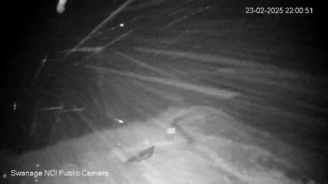 A dimly lit scene captured by a webcam, showing a snowy landscape with blurred white streaks, likely indicating falling snow or wind. The background is mostly dark, with some faint outlines of structures or obstacles. A timestamp in the corner reads 23-02-2025 22:00:51, and there is a label at the bottom left indicating "Swanage NCI Public Camera."