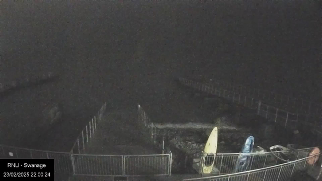 A dark scene captured by a webcam, showing a fenced area near a body of water. In the foreground, a light-colored surfboard and a blue kayak are visible on the ground. The background is mostly dark, with an indistinct shoreline and a faint outline of fencing and structures. The timestamp at the bottom indicates the image was taken on February 23, 2025, at 10:00 PM.