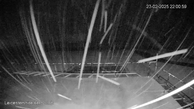 An image captured from a webcam shows a dark scene with heavy snowfall. White streaks of snow are falling diagonally across the frame, creating a blurred effect. In the background, a flat surface is partially visible, suggesting the ground covered in snow. There is a structured railing or platform edge that runs horizontally near the bottom of the image. The overall lighting is low due to the nighttime setting, and a timestamp in the corner indicates the date and time the image was taken.