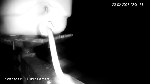 A dark image featuring a blurred white object, possibly a water tap or outlet, with a white tube extending from it. The background is mostly black, indicating low light conditions. The bottom of the image contains the text "Swanage NCI Public Camera" and a timestamp displaying the date and time.