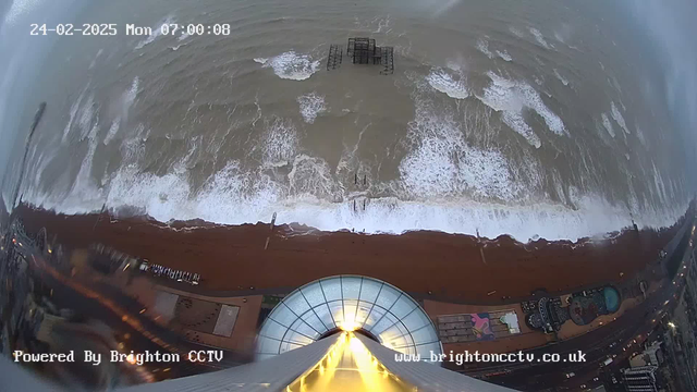 A view from a high vantage point overlooking a beach with waves crashing onto a sandy shoreline. The camera captures the ocean, which appears choppy, with white frothy waves. In the distance, a partially submerged pier structure is visible. Along the beach, there are recreational amenities and a pathway lined with lights, hinting at nearby buildings. The time displayed reads 07:00:08 on February 24, 2025.