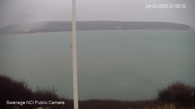 A view of a cloudy seaside landscape. The foreground features low, dark vegetation, while the background shows a calm, grayish-blue sea with several small buoys floating on the surface. The distant coastline is partially obscured by fog. A pole stands on the left side of the image. The scene appears to be in low light conditions, indicating early morning.