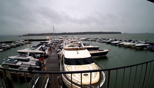 A cloudy and overcast scene of a marina with numerous boats docked. In the foreground, a large white yacht is prominently visible. Surrounding it are smaller boats of various sizes, some with blue and white stripes. The water appears choppy due to wind, and the horizon features a green hill in the distance. The atmosphere suggests a potentially stormy or rainy day.