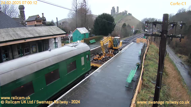 A train sits at a station with a green exterior, partially visible from the left side of the image. A yellow construction vehicle is on the platform next to the train, appearing to work on the tracks. The platform is wet, likely from rain, and there are several green benches along the platform. In the background, low hills rise with ruins of a castle at the top. Overcast skies create a gloomy atmosphere. Utility poles with wires are present on the right side of the image.