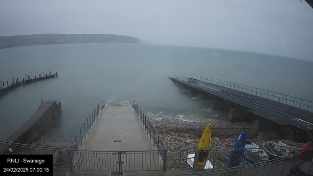 A coastal scene featuring a gray sky with low visibility due to fog. In the foreground, there is a concrete ramp leading to the water, bordered by metal railings. To the right, two boats are secured on land, with one yellow kayak visible. In the distance, a jetty extends into the water, partially submerged in the waves. The sea appears calm with small ripples. The image is timestamped at 7:00:15 on February 24, 2025.