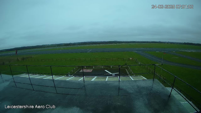 A view from a webcam showing an airport runway and surrounding grassland. In the foreground, there is a railing with a wet surface suggesting recent rain. The runway extends into the distance, with a red windsock visible to the left, indicating wind direction. The sky is overcast, and the scene appears calm and quiet. The area is mainly flat, bordered by a fence along the right side. The bottom left corner displays the text "Leicestershire Aero Club" along with a timestamp in the upper right corner.