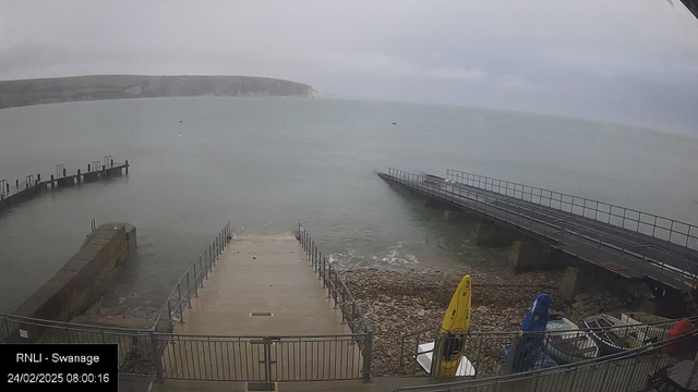 A view of a rocky shoreline with a concrete ramp leading down to the water. The water appears calm and is a muted blue-green color. In the foreground, there are two small boats on the right: one yellow and one blue. On the left, a wooden jetty extends into the water, with another jetty visible in the distance. The sky is overcast, suggesting a cloudy day, and there are some faint hills in the background.