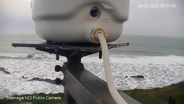 A close-up view of a white cylindrical object mounted on a metal support, with a hose attached to it. In the background, ocean waves crash against a rocky shore, and the sky appears overcast. The time and date are displayed in the corner.