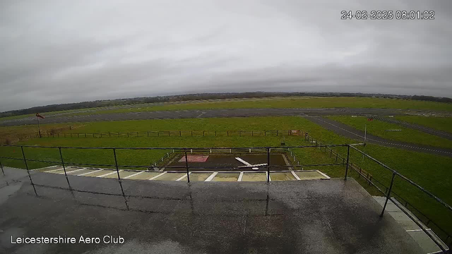 A view from a webcam looking over a mostly cloudy sky and a grassy field at the Leicestershire Aero Club. In the foreground, there is a railing and a flat surface, with a small area that appears to have been marked off or paved at the bottom. Beyond this, a wide expanse of green grass and a runway are visible, with some scattered fencing around the area. The scene is calm, with no visible aircraft or people.