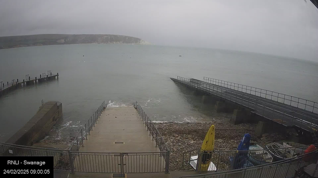 A cloudy and overcast scene depicting a shoreline with gentle waves. In the foreground, there is a concrete ramp leading down to the water, flanked by railings. To the right, there are two wooden piers extending into the water, one slightly longer than the other. On the left, there are several kayaks of various colors (yellow, blue, and red) arranged near the shore. The background features a distant view of white cliffs against a gray sky. The water is calm, and the overall atmosphere is tranquil.