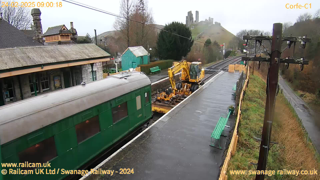 A train station scene with a green train car on the left side of the image. In front of it, a yellow construction vehicle is working on the tracks. The platform is wet, likely from recent rain, and there are several green benches along the platform. In the background, a hill with ruins can be seen, and trees are visible along the edge of the platform. The atmosphere is cloudy and overcast.