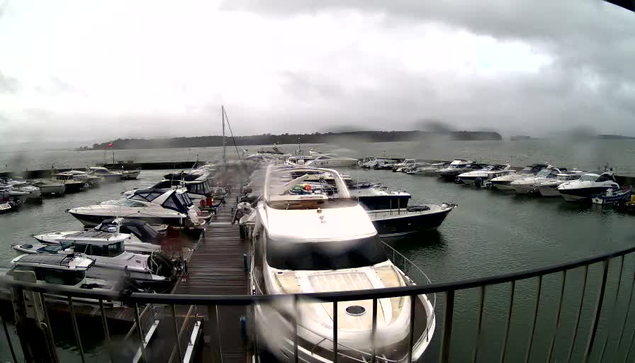 A waterfront scene depicting a marina filled with several boats of various sizes. The sky is overcast and gray, indicating possible rain. Some boats are tied to a dock made of wooden planks, while others are moored in the water. The surface of the water appears choppy, reflecting the stormy weather. In the background, a faint shoreline is visible, surrounded by trees. The image has water droplets on the lens, slightly obscuring the view.