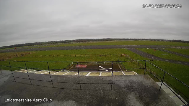 A view from a elevated balcony overlooking an empty airfield. The sky is overcast with gray clouds. In the foreground, there is a railing and a wide staircase leading down. The airfield is mostly grassy with a few runways visible, marked by thin white lines. There are scattered red boundary markers and a pentagonal area painted in red on the grass. In the background, the landscape is flat with distant trees. The image shows the time and date in the upper right corner.