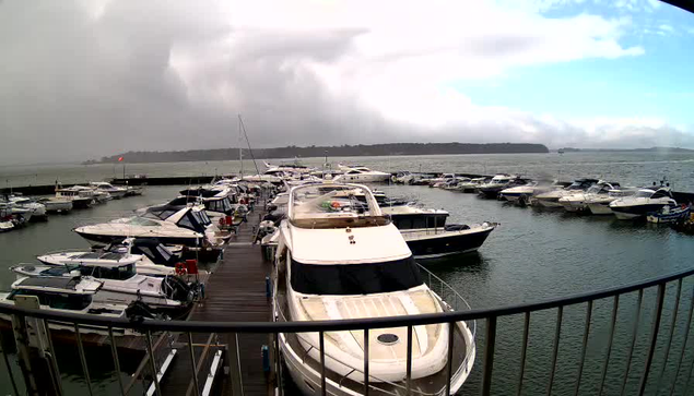 A view of a marina featuring numerous boats docked along a wooden pier. The boats vary in size and type, with some looking like yachts. The water is calm, reflecting the surroundings. In the background, there are dark clouds indicating potential rain, while the horizon has a hint of land visible. The scene conveys a quiet and somewhat overcast atmosphere.