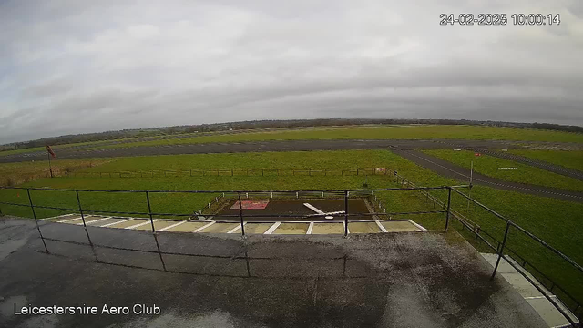 A cloudy sky with grayish tones sits above a grassy airfield. In the foreground, there is a railing from a balcony or observation deck, and the surface appears wet, possibly from recent rain. Below, the airfield is bordered by a series of fences, and there is a red and white windsock blowing gently in the breeze. A portion of the airstrip is visible, leading away into the distance where it meets more grassy areas and scattered structures. The scene conveys a quiet atmosphere typical of an airfield setting.