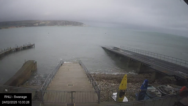 A cloudy day view of a coastal scene featuring a calm sea with gentle waves. In the foreground, a concrete boat ramp leads down to the water's edge, flanked by metal railings. To the left, a wooden jetty extends into the sea with several mooring posts visible. There are small boats gathered near the shore, including yellow, blue, and red kayaks positioned on the stony beach. The background shows cliffs and hills, partially obscured by clouds.