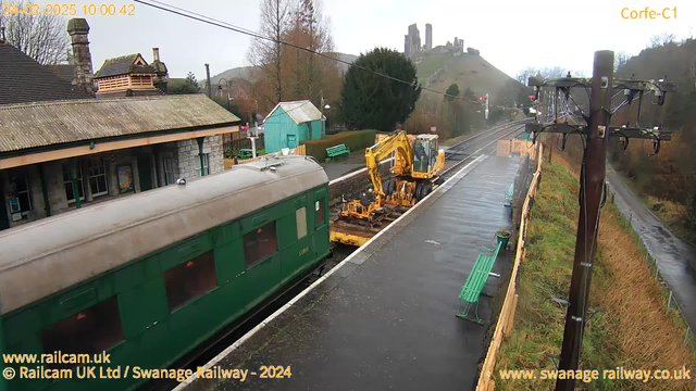 A railway station scene under overcast skies. In the foreground, a green train car is parked on the platform, beside a yellow construction vehicle performing track work. The platform is wet, likely from rain, and there are green benches along it. In the background, a hill rises with the ruins of a castle perched at the top, and trees are visible surrounding the area. Power lines and an electric pole are present on the right side of the image.