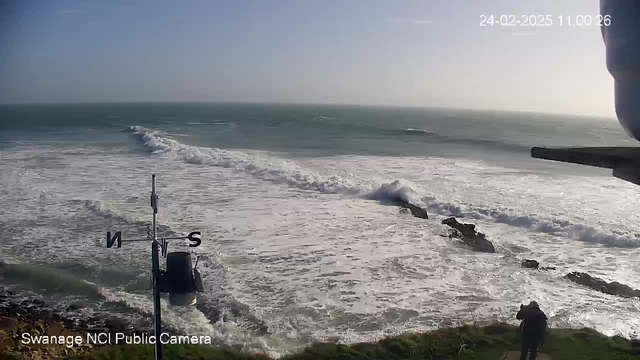 A coastal scene showing rolling waves crashing onto rocky shorelines under a clear sky. In the foreground, there is a tall weather vane with an "N" and "S" indicating north and south directions. A person is walking along a path on the right side of the image, with grass surrounding the area. The sunlight casts a warm glow over the water, highlighting the movement of the waves.