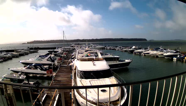 A scenic marina view featuring numerous boats in the water and docked along a wooden pier. The sky is partly cloudy with some blue visible. In the foreground, a larger boat is docked, while various smaller boats are lined up along the marina. The water appears calm and reflective, with a distant shoreline visible on the horizon.