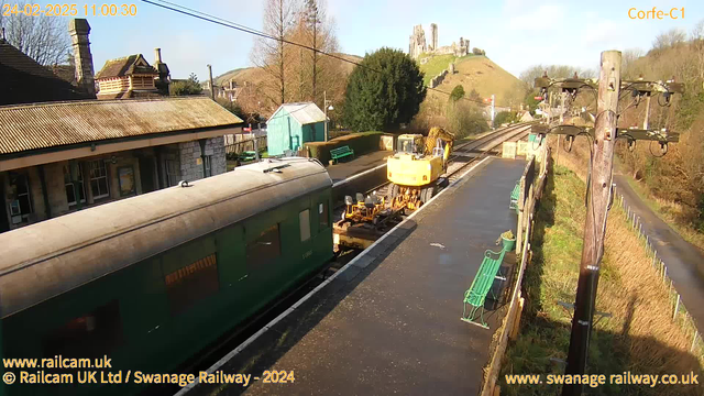 A view of a railway station platform with a green train car parked alongside. A yellow piece of construction equipment is positioned on the tracks beside the train. In the background, a stone castle ruins can be seen atop a hill, surrounded by trees. The scene is bright and sunny, reflecting clear blue skies. The platform features green benches and the edges are lined with hedges. A wooden utility pole is visible on the right side of the image.