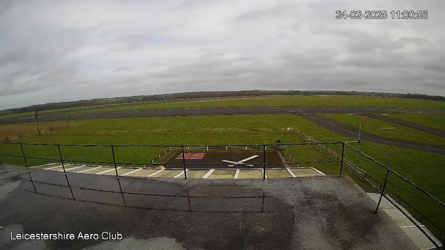 A cloudy sky is overhead, with a mostly overcast view at the Leicestershire Aero Club. Below, there is a grassy field that transitions into a runway. A white airplane silhouette is painted on the ground near the edge of a tarmacked area, with some markings indicating a landing zone. There are also red and white warning signs visible along the perimeter of the runway. The scene suggests an active aviation area, but no aircraft are present at the moment.