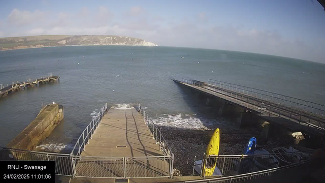 A view of a coastal scene featuring a calm sea under a clear sky with some clouds. In the foreground, a concrete ramp leads down to the water, and there is a wooden pier extending into the sea on the left side. On the right side, two kayaks in yellow and blue are positioned near the shore, alongside some scattered rocks. The distant cliffs are visible across the water, creating a scenic backdrop. The image indicates a sunny day in Swanage, with a timestamp in the corner.