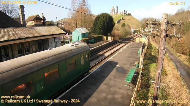 A sunny day at a railway station. A green train is parked on the left side of the image, with its windows visible. Adjacent to the train, there is a platform with several green benches. In the background, there are trees and a building with a tiled roof. A hill rises in the distance, topped with a castle or ruins. The sky is clear, and there are power lines stretching across the image. The scene conveys a quiet and picturesque setting.