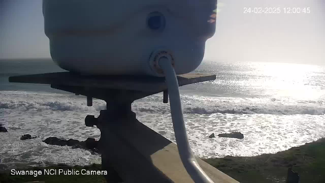 A webcam view captures a close-up of a white cylindrical object mounted on a platform, with a white cable extending from it. In the background, a bright ocean scene is visible, with sunlight reflecting off the water and waves crashing against dark rocks near the shore. The image is taken on February 24, 2025, at noon, and text at the bottom indicates "Swanage NCI Public Camera."