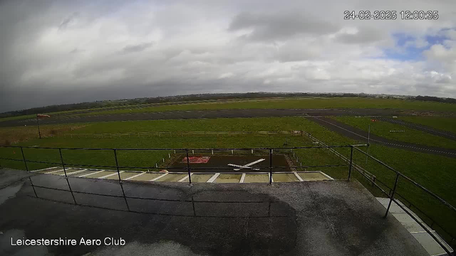 A view from a webcam overlooking a grassy airfield under a cloudy sky. In the foreground, there is a rooftop ledge with railings. Below, a large grassy area is visible with a red and white windsock indicating wind direction. A small airstrip runs through the center, leading to a flat, paved surface. Several small aircraft parking areas can be seen in the distance, surrounded by green fields and low wooden fencing. The setting appears calm with no visible aircraft in the immediate vicinity.